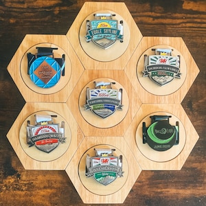 May include: A wooden display with seven medals in circular holders set in a hexagonal frame. The medals, with text and graphics, commemorate events like "EDALE SHYLINE" and "WELSH 3000S". The medals are silver and feature various colors.