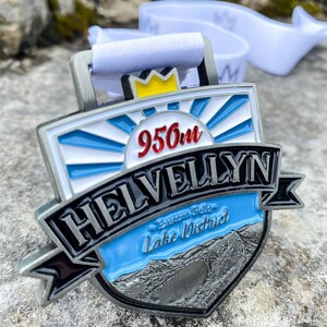 Op de afbeelding: Een zilveren medaille met een blauw, wit en zwart ontwerp. Het bevat de tekst "Helvellyn" en "956m" met een kroon erboven. De tekst "Eastern Fells Lake District" is ook inbegrepen.