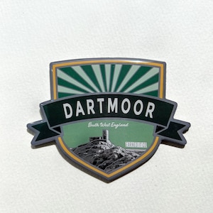 May include: A green and grey shield-shaped sticker with the text "Dartmoor" in white letters. The sticker also has the text "South West England" and "Earned It Co" in smaller white letters. The sticker has a green and white sunburst design in the background and a black and white illustration of a castle on a hill.