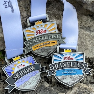 Pode incluir: Três medalhas de prata com detalhes azuis, amarelos e roxos. Cada medalha apresenta uma cordilheira e o nome da montanha, juntamente com a altura em metros. As medalhas dizem "Scafell Pike - Lake District 978m", "Helvellyn - Eastern Fells - Lake District 950m" e "Skiddaw - Lake District 931m".