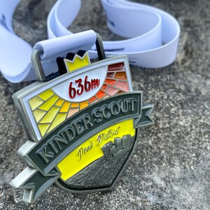 Op de afbeelding: Een zilveren medaille met een geel, rood en oranje glas-in-loodontwerp. De medaille heeft de tekst "Kinder Scout Peak District 636m".