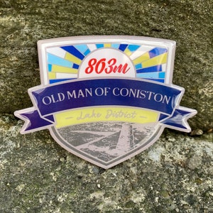 May include: A blue and yellow enamel pin with a white background. The pin features a stylised image of a mountain range with the text "Old Man of Coniston - Lake District - 863m".