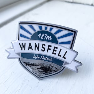 May include: A silver and blue enamel pin with a white banner that reads "Wansfell Lake District" and a blue circle with the text "487m".