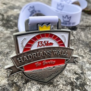 May include: A silver medal with a red and white design. The medal features the text "Hadrian's Wall The Borders" and "135km".