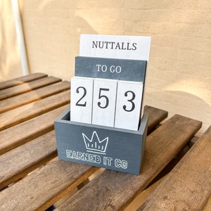 May include: A grey wooden box with a white crown and the text "Earned It Co." on the front. The box holds three white wooden blocks with the numbers 2, 5, and 3 on them. The blocks are stacked on top of two grey wooden blocks with the text "Nuttalls" and "To Go" on them.