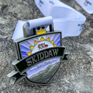 May include: A silver medal with a black and white design. The medal features a stylized sun with a purple and blue background, the text "SKIDDAW" in black, and the text "Lake District" in white. The medal also includes the text "931m" in red.