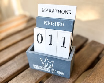 Może przedstawiać: Drewniany kalendarz blokowy ze słowami "MARATHONS" i "FINISHED" wydrukowanymi na górnych blokach. Dolne bloki wyświetlają liczby "011". Kalendarz znajduje się w szarej drewnianej skrzynce z napisem "EARNED IT CO" i grafiką korony.