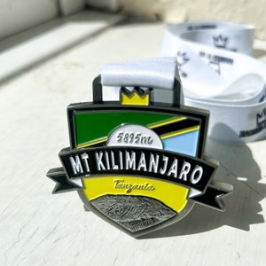 Puede incluir: Una medalla de plata con un diseño de escudo verde, amarillo y azul. El escudo presenta el texto "MT KILIMANJARO" y "Tanzania" con una imagen de cordillera. La medalla tiene 5895 m de altura.