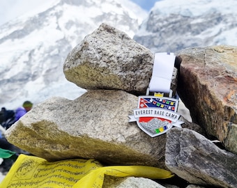 May include: A silver medal with a colourful shield design and the text "Everest Base Camp". The medal is resting on a pile of rocks with a snowy mountain in the background.