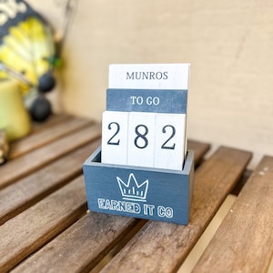 May include: A wooden countdown block with the numbers 282, the words "MUNROS", "TO GO", and "EARNED IT CO" in white lettering on a dark blue background.