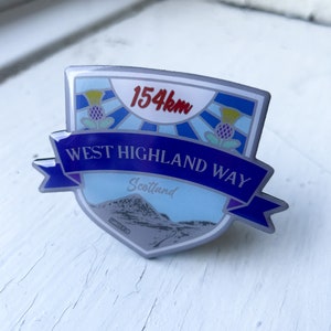 May include: A blue and silver enamel pin with a white shield design. The shield features a mountain range, a road, and the text "Scotland". The top of the shield has a blue and white striped design with the text "154km" and a yellow and green thistle. The bottom of the shield has a blue ribbon with the text "West Highland Way".
