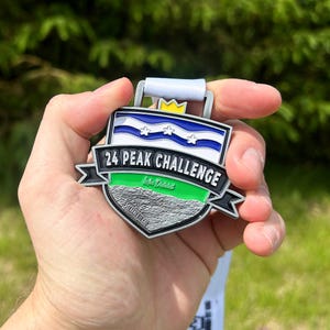 May include: A silver-colored medal with a white ribbon. The medal has a shield shape with the text "24 PEAK CHALLENGE" and a blue and white design. The medal also has a green stripe and a gold crown.