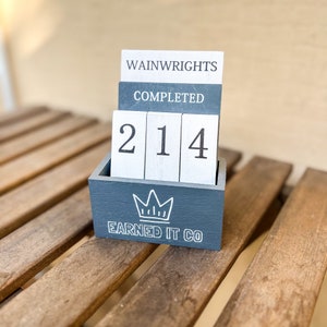 May include: A gray wooden box with a crown design and the text "Earned It Co."  The box holds four white blocks with the numbers "214" and the text "Wainwrights" and "Completed" on the top two blocks.