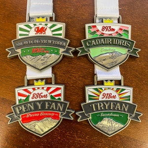 Puede incluir: Cuatro medallones de plata con diseños coloridos y escenas de montaña. Cada medalla tiene el nombre de una montaña, su altura en metros y la región en la que se encuentra. Las medallas son Snowdon Yr Wyddfa, 1085m, Cadair Idris, 893m, Pen Y Fan, 886m, y Tryfan, 918m.
