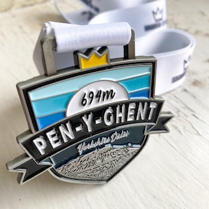 May include: A silver medal with a blue, white and black design. The medal features the text "PEN-Y-GHENT" and "694m" and a graphic of a mountain range. The text "Yorkshire Dales" is also included on the medal.