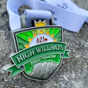 Op de afbeelding: Een groene en zilveren medaille met de woorden "High Willhays" en "Dartmoor National Park" erop. De medaille heeft ook een zon met de tekst "621m" erop.