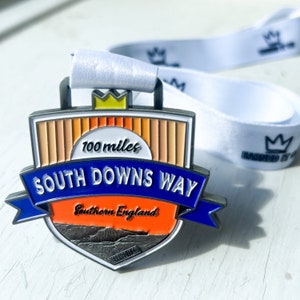 May include: A silver medal with a blue and orange design. The medal features the text "100 miles South Downs Way Southern England".