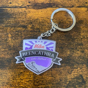 May include: A purple and silver keychain with the text "868m Blencathra - Lake District -" on it. The keychain is shaped like a shield with a mountain peak in the background.