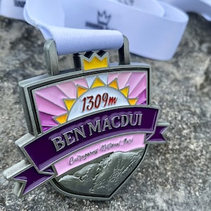 May include: A silver medal with a purple, pink, and yellow design. It features a stylized sun with the text "1309m BEN MACDUI Cairngorms National Park".