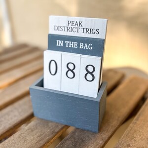 Op de afbeelding: Een grijze houten doos met een schuifregelaar die het nummer 088 weergeeft. De doos heeft een wit label met de tekst "Peak District Trigs" en "In The Bag".