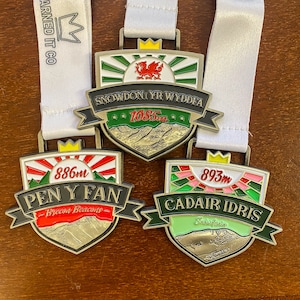 Könnte beinhalten: Drei Metallmedaillen mit weißen Bändern. Jede Medaille zeigt ein Schilddesign mit einer Berglandschaft und dem Namen eines Berges. Die Medaillen sind mit "Snowdon Yr Wyddfa", "Pen Y Fan" und "Cadair Idris" beschriftet. Snowdon ist 1085m, Pen Y Fan ist 886m und Cadair Idris ist 893m.