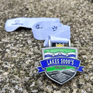 May include: A silver medal with a blue, green, and white design. It says "Lakes 3000's" and "83KM, 4000M".