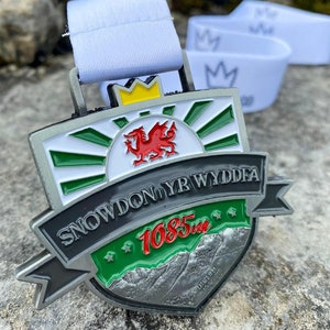 Puede incluir: Una medalla de plata con un diseño verde, blanco y rojo. La medalla presenta un dragón rojo, el texto "SNOWDON YR WYDDFA" y el número "1685" en rojo. La medalla está colgada de una cinta blanca.