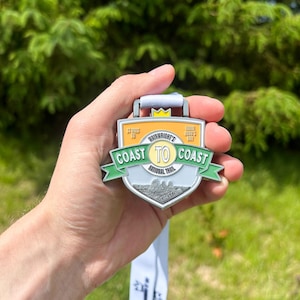 Może przedstawiać: Medal Coast to Coast National Trail ze srebrnym, zielonym i złotym wzorem. Medal ma napis "Coast to Coast" i "Wainwright's". Medal jest trzymany przez osobę.