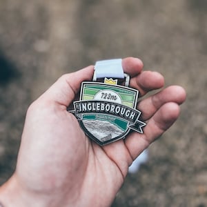 May include: A silver medal with a green and white design. The medal features the text "723m Ingleborough Yorkshire Dales".