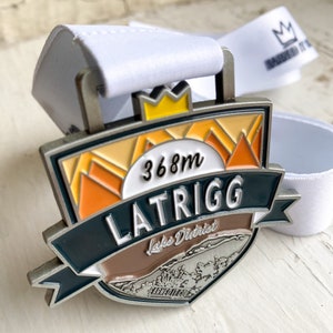 May include: A silver medal with a blue, orange, and brown design. The medal features the text "Latrigg Lakes District 368m" and a crown.
