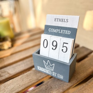 May include: Grey wooden box with a white crown and the text "Earned It Co." on the front. The box holds white wooden blocks with the numbers "095" and the text "Ethels Completed" on the top.