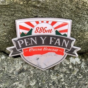 May include: A red, white and green enamel pin with a mountain range design. The pin reads "886m PEN Y FAN - Brecon Beacons -"