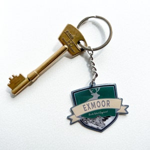 May include: A gold-colored key and a keychain with an Exmoor design. The keychain has a green and beige shield shape with a deer silhouette and the word "EXMOOR". The key is next to the keychain.