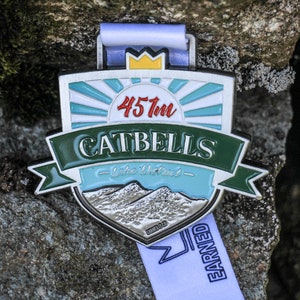 May include: A silver medal with a green and blue design. The medal features a sunrise with the text "457m Catbells - Lake District - Earned".