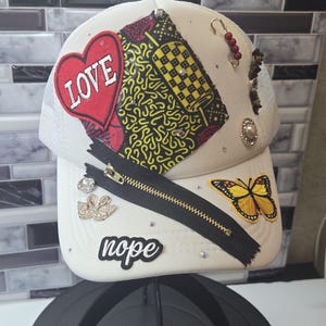 Pode incluir: Boné de caminhoneiro branco com parte traseira em rede e aba curva. O boné é decorado com um coração vermelho com a palavra "LOVE", um zíper preto, uma borboleta amarela e a palavra "nope". Inclui strass e patches de tecido.