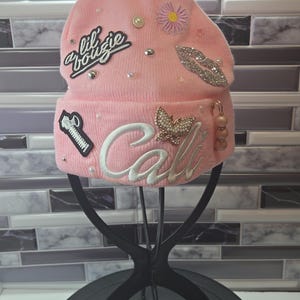 May include: A pink knitted beanie hat adorned with various embellishments. The hat displays the text "lil bougie" and "Cal" in cursive, alongside a butterfly, a zipper, a flower, and lips. It is further decorated with pearls and rhinestones.