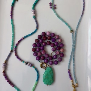 May include: Two long beaded necklaces and one shorter beaded necklace with a large green stone pendant. The necklaces feature a variety of small, round, faceted, and smooth beads in shades of purple, teal, light blue, and lavender with gold-tone accents.