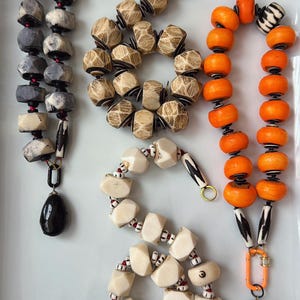 May include: Four necklaces arranged on a white surface. One features grey and white faceted beads with a black pendant. Another is a tan wood-textured bead strand. A third has bright orange beads with a black and white patterned pendant. One is cream-colored.