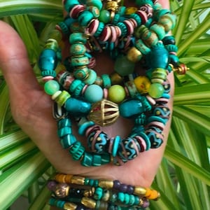 May include: A collection of colorful beaded bracelets and necklaces. The jewelry features turquoise, green, pink, and gold beads in various shapes and sizes. Some bracelets have gold accents and purple beads. The jewelry is displayed against a backdrop of green foliage.