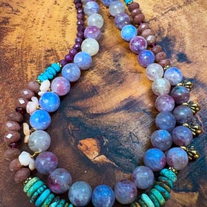 May include: A multi-strand necklace featuring large, round beads in shades of blue, purple, and white, accented with turquoise and smaller, faceted beads. The necklace has a gold clasp and is displayed on a wooden surface.