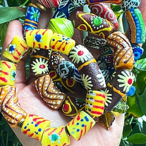 May include: A colorful beaded bracelet with a variety of patterns and colors, including yellow, brown, red, and blue. The beads are cylindrical and appear to be made of painted clay or ceramic. The bracelet is held in a hand.
