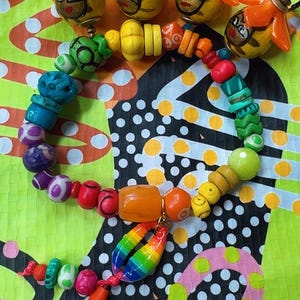May include: A colorful necklace with a variety of beads in shades of blue, green, yellow, orange, and red. The necklace also features several pendant charms with painted faces and a rainbow-colored charm. The background is a patterned fabric.