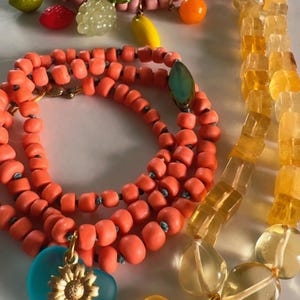 May include: A collection of colorful beaded necklaces. One necklace is pink with fruit charms, another is orange, and a third is yellow. The orange necklace has a blue heart pendant with a gold sunflower. The yellow necklace has a yellow heart pendant with a gold sunflower.