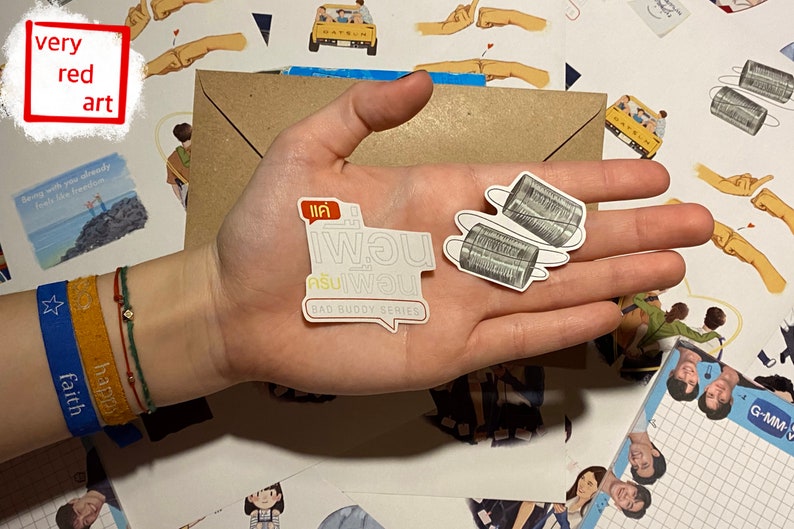 Op de afbeelding: Twee witte stickers met zwarte tekst en afbeeldingen. De eerste sticker zegt "แค่ พอน" en "Bad Buddy Series" in het Engels. De tweede sticker toont twee blikken die met een touwtje aan elkaar zijn verbonden.