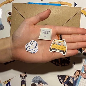 Op de afbeelding: Een hand die een aantal stickers vasthoudt, waaronder een blauw recycle-symbool, een witte sticker met een smiley en Thaise tekst, en een gele Datsun-auto sticker met mensen erop.