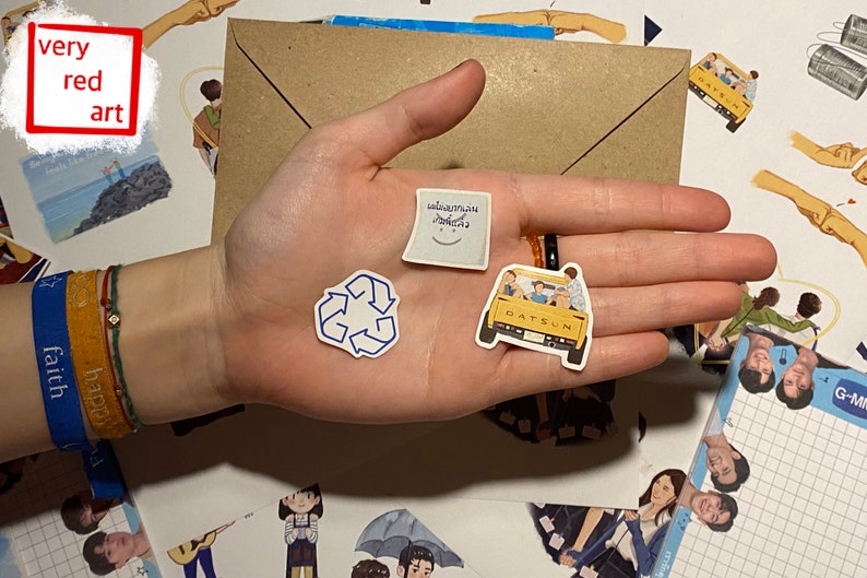 Op de afbeelding: Een hand die een aantal stickers vasthoudt, waaronder een blauw recycle-symbool, een witte sticker met een smiley en Thaise tekst, en een gele Datsun-auto sticker met mensen erop.