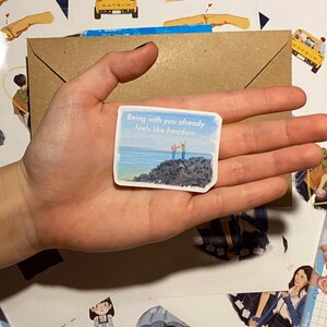 Op de afbeelding: Een hand die een klein kaartje vasthoudt met de tekst "Met jou zijn voelt al aan als vrijheid" erop gedrukt. Het kaartje heeft een aquarel achtergrond met een blauwe lucht, zee en twee figuren die op een rotsachtige kust staan.