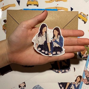 Op de afbeelding: Een sticker met twee vrouwen in schooluniform die elkaar vasthouden. De sticker is wit met een lichtblauwe achtergrond en heeft een lichtblauwe rand. De vrouwen glimlachen en kijken elkaar aan. De sticker zit op een bruine envelop met andere stickers eromheen.