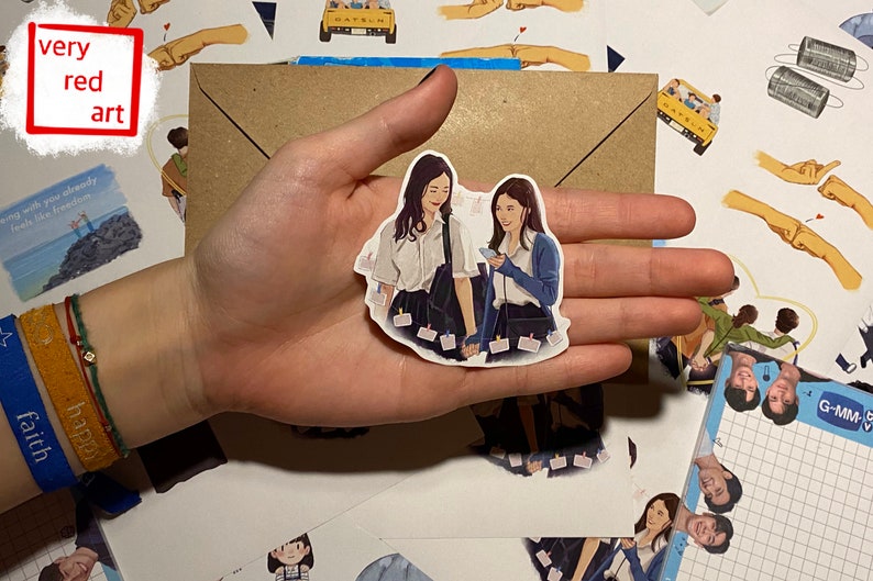 Op de afbeelding: Een sticker met twee vrouwen in schooluniform die elkaar vasthouden. De sticker is wit met een lichtblauwe achtergrond en heeft een lichtblauwe rand. De vrouwen glimlachen en kijken elkaar aan. De sticker zit op een bruine envelop met andere stickers eromheen.