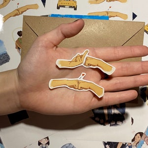 Op de afbeelding: Twee gele handgetekende stickers van twee mensen die een pinky promise doen. De stickers zitten op een hand met een blauwe en gele armband.